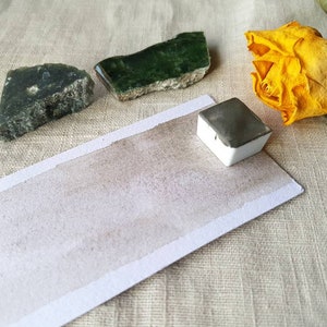 May include: A watercolor pan with a gray paint sample on a white paper card. The card is on a beige linen fabric background. There are two green stones and a dried yellow rose on the fabric.