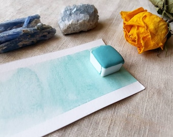 Può includere: Un blocco di pittura ad acquerello turchese su un campione dipinto su carta bianca. L'immagine include anche cristalli blu e una rosa gialla secca, suggerendo un tema di arte ed elementi naturali.
