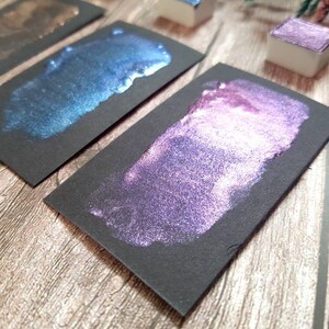 May include: Three black cardstock swatches with metallic paint samples. The swatches show a dark brown, a teal blue, and a purple with a pink shimmer.