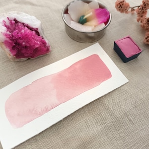 Pode incluir: Uma amostra de tinta aquarela rosa em um cartão branco. A tinta é de cor rosa claro e tem um efeito degradê. O cartão está sobre uma superfície de tecido bege.
