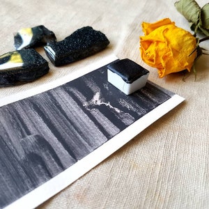 Pode incluir: Uma pastilha de aquarela preta com um rótulo branco, uma rosa amarela e um pedaço de papel estampado em preto e branco. O papel tem uma lavagem de aquarela preta.