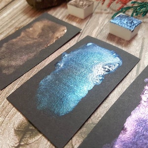 May include: Three samples of metallic watercolor paint on black paper. The colors are a shimmery bronze, a teal blue with silver flecks, and a purple with silver flecks.