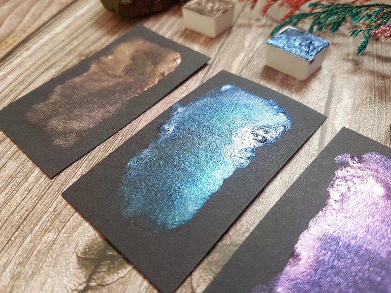 May include: Three samples of metallic watercolor paint on black paper. The colors are a shimmery bronze, a teal blue with silver flecks, and a purple with silver flecks.