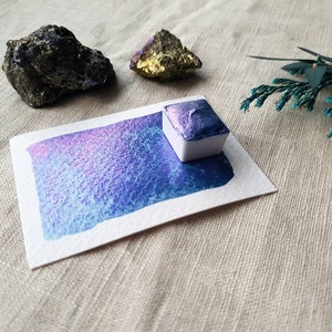 Pode incluir: Uma pequena bandeja quadrada branca de tinta aquarela azul iridescente com uma amostra de pincelada em um cartão branco. A bandeja está sobre uma superfície de tecido bege.