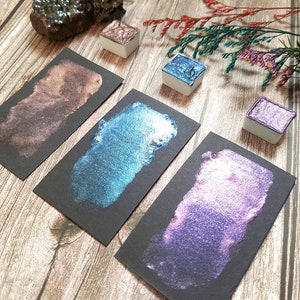 May include: Four metallic watercolor paint pans on black paper swatches. The colors are a deep brown, a teal blue, a purple, and a light pink.