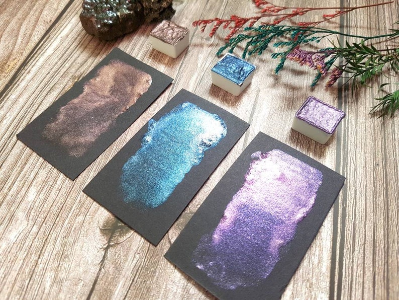 May include: Four metallic watercolor paint pans on black paper swatches. The colors are a deep brown, a teal blue, a purple, and a light pink.