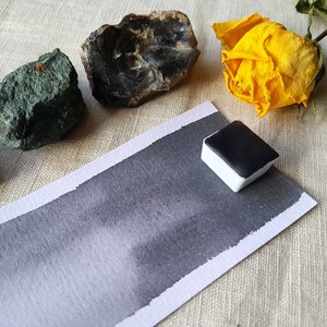 May include: A black watercolor paint pan with a sample of the paint on a white paper card. The card is on a beige linen fabric background. There are three rocks and a dried yellow rose on the fabric.