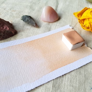 May include: A watercolor pan with a light peach color, sitting on a white paper with a gradient of peach color. The paper is on a beige fabric surface. There are other objects in the background, including a pink stone, a gray stone, and a yellow flower.