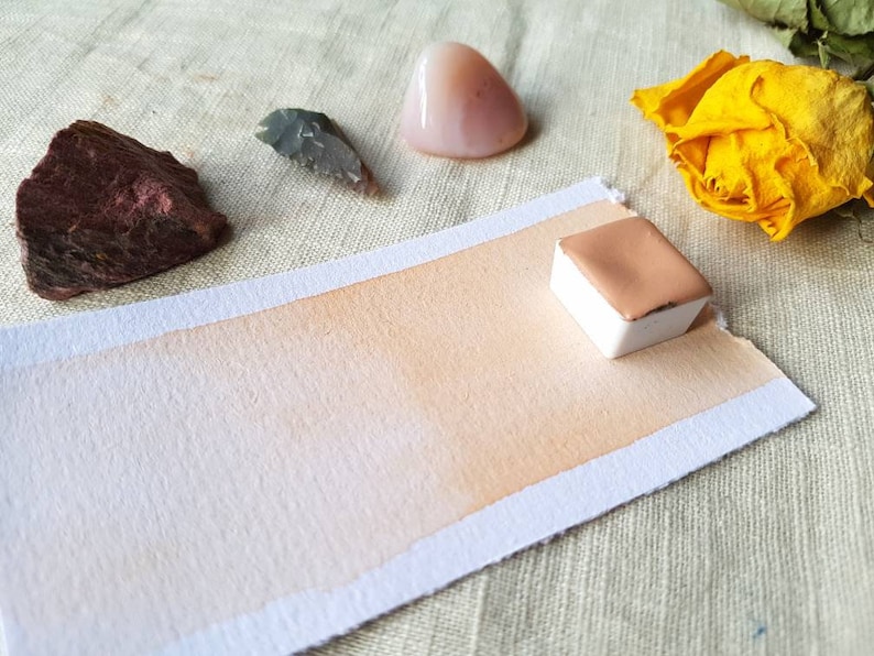 May include: A watercolor pan with a light peach color, sitting on a white paper with a gradient of peach color. The paper is on a beige fabric surface. There are other objects in the background, including a pink stone, a gray stone, and a yellow flower.