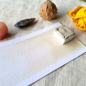 May include: A white watercolour pan with a light brown watercolour wash on a white paper surface. The paper is on a beige linen fabric background. There are other objects in the image, including a pink stone, a brown stone, and a dried yellow rose.