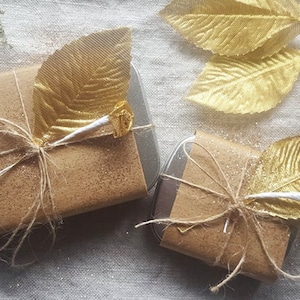 Pode incluir: Duas pequenas caixas de presente embrulhadas em papel castanho com glitter dourado e amarradas com barbante. Cada caixa é decorada com uma folha dourada e uma fita branca.