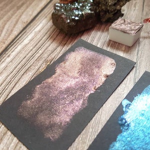 May include: Two samples of metallic watercolor paint on black paper. One sample is a shimmery rose gold color, the other is a shimmery teal blue.