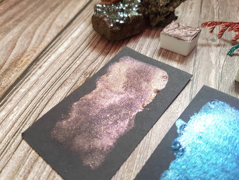 May include: Two samples of metallic watercolor paint on black paper. One sample is a shimmery rose gold color, the other is a shimmery teal blue.
