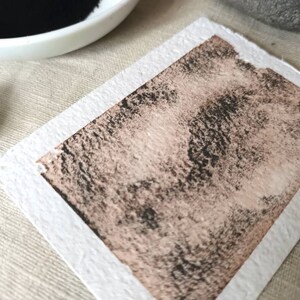 May include: A close-up of a textured, light brown paper with a dark gray wash of paint applied to it. The paper has a white border.