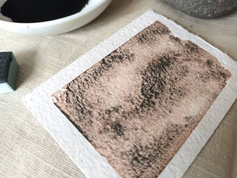 May include: A close-up of a textured, light brown paper with a dark gray wash of paint applied to it. The paper has a white border.