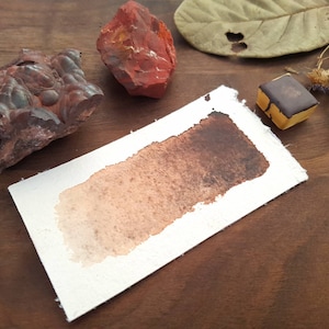Puede incluir: Un primer plano de una acuarela lavada sobre un fondo de papel blanco. El lavado es un degradado de colores marrón y naranja. El papel está sobre una superficie de madera.
