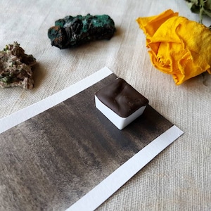 May include: A watercolor pan with a dark brown paint sample on a white card. The card is on a white linen fabric background. There are three small rocks on the fabric, one green, one brown, and one green and brown.