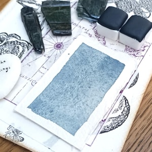 Könnte beinhalten: Eine Ansammlung von Kunstbedarf und dekorativen Gegenständen auf einer Holzoberfläche. Enthält Aquarellfarbe, einen herzförmigen Stein mit dem Text "enjoy the little things" und kleine, dunkel gefärbte Steine. Ein kartenähnliches Design ist im Hintergrund zu sehen.