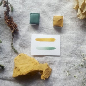 Pu&ograve; includere: Due pastiglie di acquerello, una verde e una gialla, con un cartoncino bianco che mostra un lavaggio ad acquerello verde e giallo. Il cartoncino &egrave; su uno sfondo di lino bianco con fiori secchi e una roccia gialla.