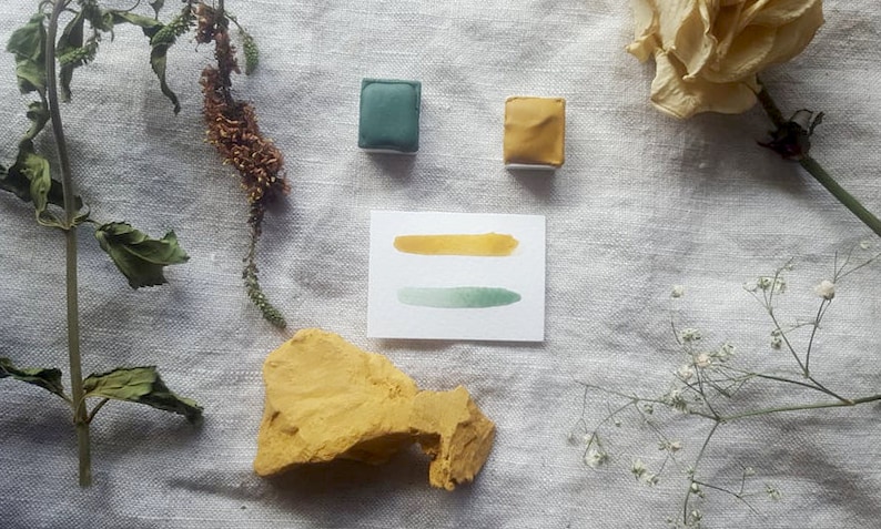 Pu&ograve; includere: Due pastiglie di acquerello, una verde e una gialla, con un cartoncino bianco che mostra un lavaggio ad acquerello verde e giallo. Il cartoncino &egrave; su uno sfondo di lino bianco con fiori secchi e una roccia gialla.