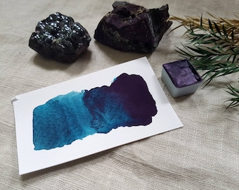 Può includere: Una tavolozza di acquerello con un campione di vernice verde acqua e viola su un cartoncino bianco. La tavolozza di vernice è di colore blu chiaro con un coperchio viola. Il campione di vernice è un gradiente dal verde acqua al viola. Sullo sfondo ci sono due pietre grigie scure e un ramoscello di fogliame verde.