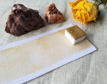 May include: A still-life composition featuring a watercolor paint swatch, a small paint pan, two rocks, and a yellow rose. The paint swatch is a light brown color, and the paint pan is white with a gold top. The rocks are brown and tan.