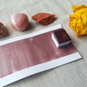 K&ouml;nnte beinhalten: Nahaufnahme einer Aquarellfarbpalette mit einem dunkelbraunen Pigment. Die Palette liegt auf einer wei&szlig;en Karte mit einem braunen Aquarell-Wasch. Im Hintergrund befinden sich drei kleine Steine und eine gelbe Blume.