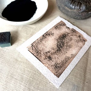 May include: A close-up of a black watercolor pan and a small square of black watercolor paint on a white surface. The paint has been applied to a piece of white paper, creating a dark brown, textured wash.