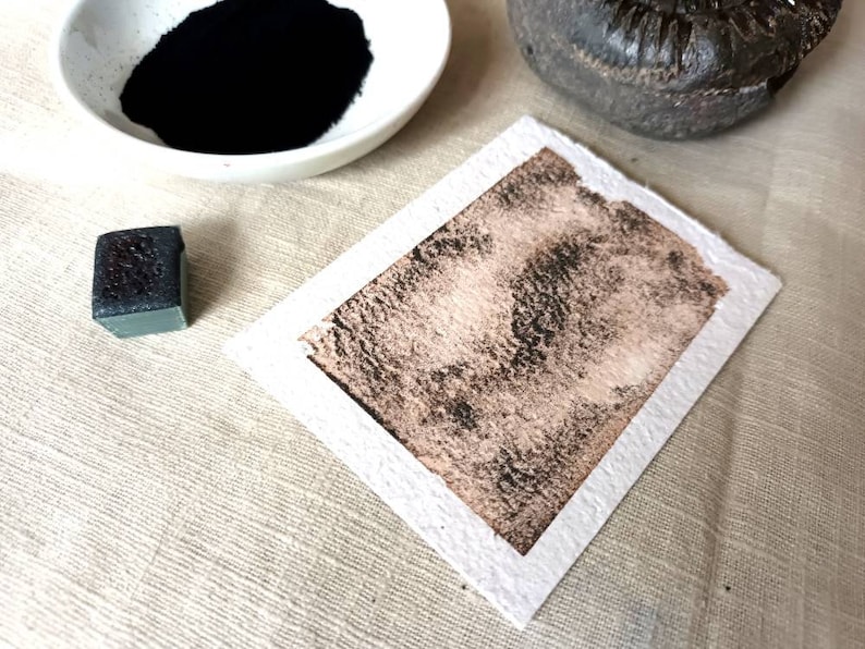 May include: A close-up of a black watercolor pan and a small square of black watercolor paint on a white surface. The paint has been applied to a piece of white paper, creating a dark brown, textured wash.
