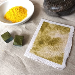 Puede incluir: Dos pequeñas pastillas de pintura acuarela verde sobre una superficie de lino blanco. Un pequeño cuenco blanco con polvo amarillo se encuentra en el fondo. Una lavada de acuarela verde es visible en un trozo de papel.
