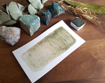 May include: A watercolor paint pan with a green pigment swatch on a white paper card. The card is on a wooden surface with various rocks and greenery.