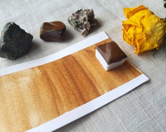 May include: A watercolor paint pan with a brown pigment. The pan is sitting on a white paper with a brown watercolor wash. There are also a few small rocks and a yellow rose on the table.