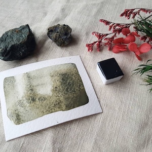 Könnte beinhalten: Ein Aquarellbild einer grünen und weißen Landschaft auf einer weißen Karte. Eine kleine schwarze Aquarellschale und zwei dunkelgraue Steine liegen auf einer weißen Leinenoberfläche. Im Hintergrund befindet sich ein Zweig mit roten Blüten und grünen Blättern.