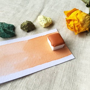 May include: A watercolor paint pan with a light orange color swatch on a white paper background. The paint pan is a square shape and is made of white plastic. The paint is a light brown color.