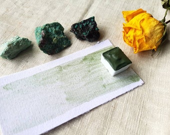 May include: A green watercolor paint pan with a green paint swatch on a white paper card. There are three green rocks and a dried yellow rose on a beige fabric background.