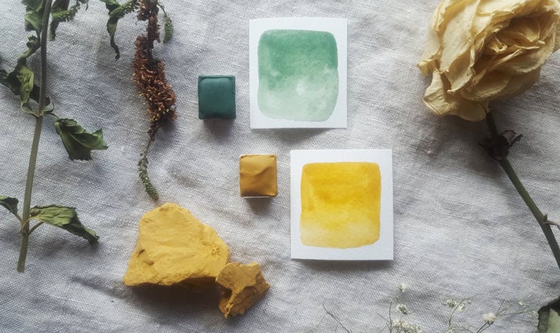 Pu&ograve; includere: Una vista dall'alto di acquerelli, un fiore secco e una roccia. Gli acquerelli sono in piccole vaschette quadrate. I colori sono verde e giallo. Il fiore secco &egrave; una rosa bianca. La roccia &egrave; di colore marrone chiaro.