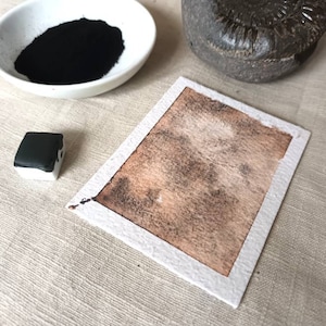 Puede incluir: Una acuarela marrón sobre un fondo de papel blanco. La acuarela es desigual y tiene un aspecto moteado. Hay una pequeña pastilla de acuarela negra y un cuenco blanco con polvo de acuarela negra en el fondo.