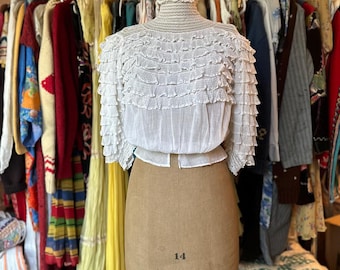 May include: A white, ruffled blouse displayed on a vintage dress form. The blouse has a high, ruffled collar and layers of delicate ruffles across the bodice and sleeves. The dress form is tan and has the number "14" and "MODEL 1945" printed on it. A rack of vintage clothing is in the background.