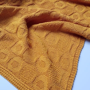 Puede incluir: Una manta de punto de color amarillo mostaza con un patrón texturizado. La manta está doblada y drapeada sobre una superficie blanca.