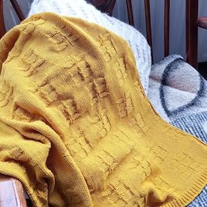 Puede incluir: Manta de punto amarillo mostaza con un patrón geométrico sobre una silla de madera. La silla tiene un acabado marrón oscuro y un respaldo decorativo. Se ven otras mantas y almohadas.