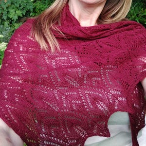 Puede incluir: Un chal de punto color burdeos con un delicado patrón de encaje. El chal está drapeado sobre los hombros de una persona y está hecho de un hilo suave y ligero.