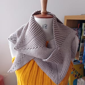 Puede incluir: Un chal gris tejido con un diseño geométrico está drapeado sobre un maniquí. El chal tiene una textura acanalada y se muestra sobre una parte superior amarilla acanalada. El maniquí está sobre un soporte de madera.