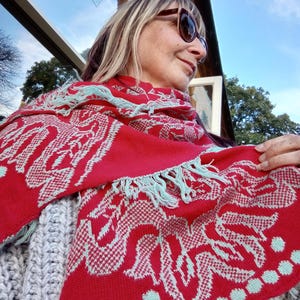 Puede incluir: Una bufanda de punto roja con un patrón geométrico blanco y azul claro. La bufanda tiene flecos en los extremos.