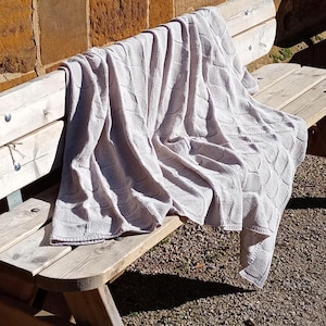 Puede incluir: Una manta de tejido de punto beige claro con un patrón texturizado está sobre un banco de madera. El diseño de la manta presenta un patrón geométrico repetitivo. El banco es de madera clara y está al aire libre.