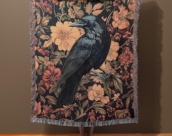 Può includere: Un corvo nero con un riflesso blu si posa su un ramo di un arazzo floreale. L'arazzo presenta una varietà di fiori in tonalità di rosa, giallo e rosso, con foglie e viti verdi.