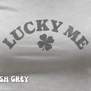Close-up of the women's baby tee crop top in ash grey. The shirt has a distressed design of a four-leaf clover and says, "Lucky Me."