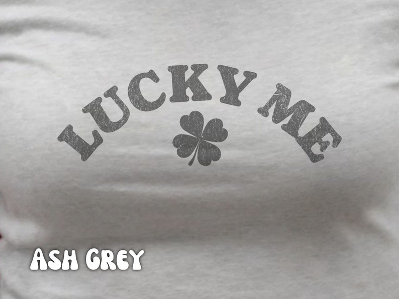Close-up of the women's baby tee crop top in ash grey. The shirt has a distressed design of a four-leaf clover and says, "Lucky Me."
