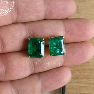 Könnte beinhalten: Ein Paar Ohrstecker mit smaragdgrünen Edelsteinen im Smaragdschliff, die jeweils mit vier kleinen goldenen Krappen gefasst sind. Die Ohrringe sind rechteckig und haben eine facettierte Oberfläche. Der Text THE GLAM FACTORY JEWELS ist sichtbar.