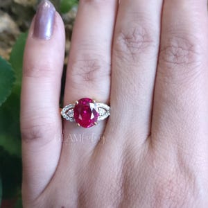 May include: A gold ring featuring a large oval ruby gemstone, flanked by smaller white stones. The ring is displayed on a hand with painted nails. The text "the GLAMfactory" is visible in the background.