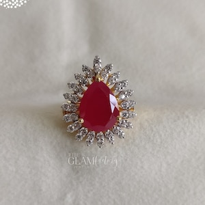 以下が含まれることがあります： 大きな洋梨型の赤い宝石が、小さな透明な宝石で囲まれた、金の指輪です。